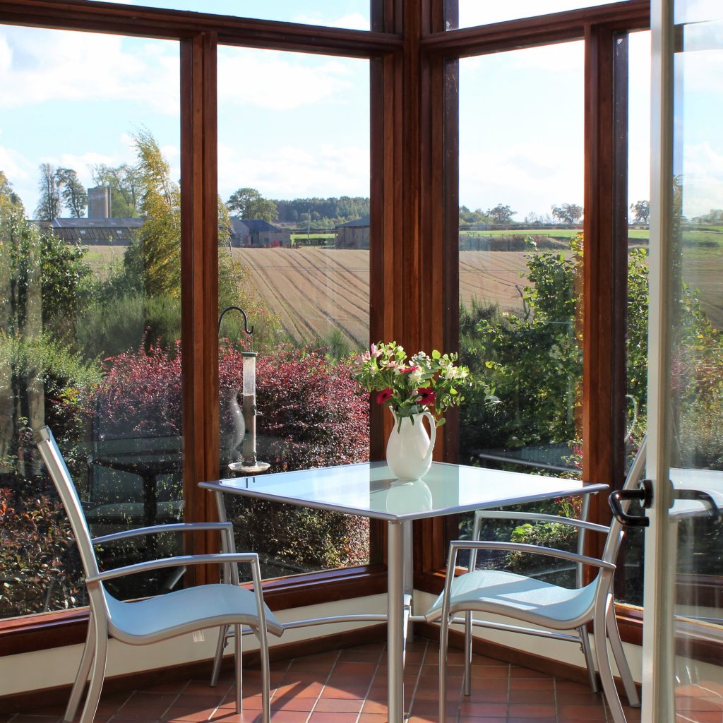 Vase of flowers on a table in a sunny conservatory looking out over the cottage garden to fields beyond
